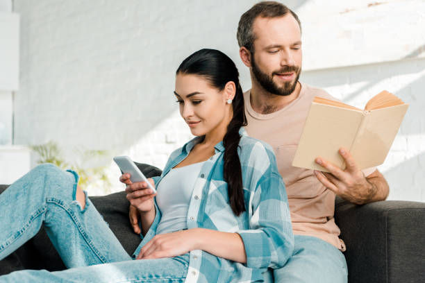 istock beautiful wife using smartphone while man sitting on couch and reading book at home 1071052178