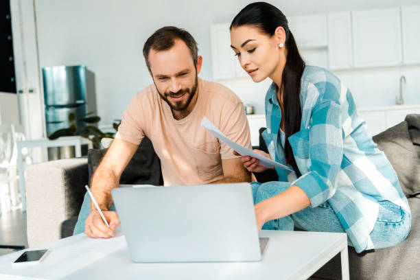 istock couple sitting on couch, filling paperwork and using laptop at home 1071052206