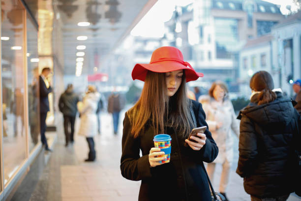 fille marche et le langage sms sur le téléphone intelligent dans la rue, coiffé d’un chapeau rouge - red text messaging cafe teenage girls photos et images de collection