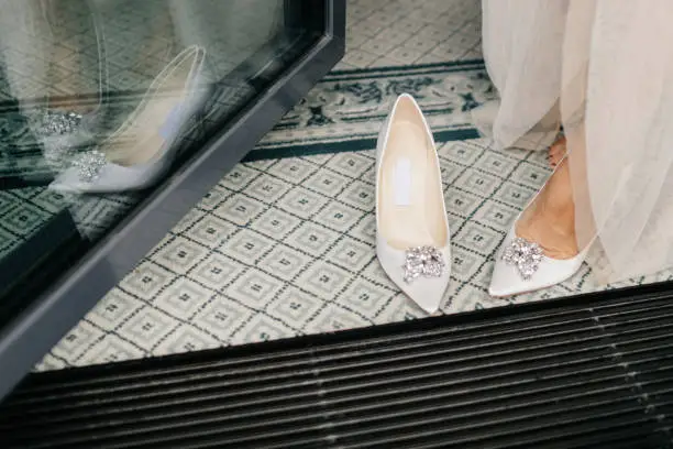 Photo of Unrecognizable bride in white dress, puts on wedding shoes, has tender feet, preparares for special occasion in her life. Brides white luxury shoes