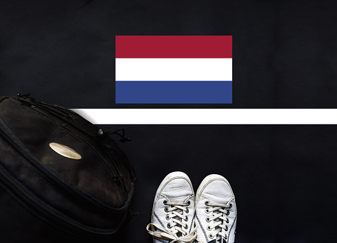 a man with a shoes and backpack is standing on asphalt next to flag of Netherlands and border.