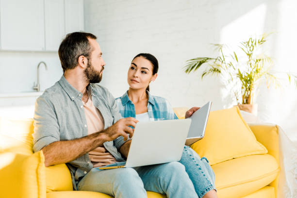 istock handsome husband using laptop while beautiful wife holding notebook and sitting on couch 1071072112