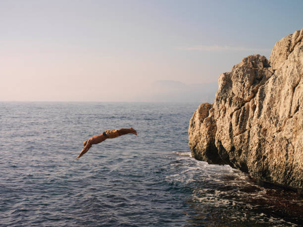 Man dives off cliff into sea He is mid air and in diving formation cliff jumping stock pictures, royalty-free photos & images