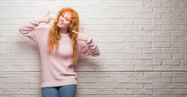 istock Young redhead woman standing over brick wall smiling confident showing and pointing with fingers teeth and mouth. Health concept. 1071094548