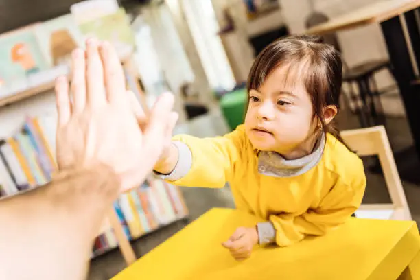 High five. Dark-eyed sunny child giving high five to her teacher in the inclusive kindergarten