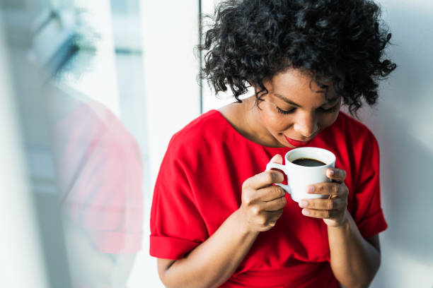 un primer plano de una mujer de pie junto a la ventana con una taza de café. - young adult lifestyles city life drinking fotografías e imágenes de stock