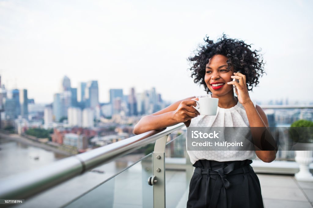 A businesswoman standing against London view panorama, making a phone call. A portrait of black businesswoman with a cup of coffee standing against London rooftop view panorama, making a phone call. Copy space. Businesswoman Stock Photo