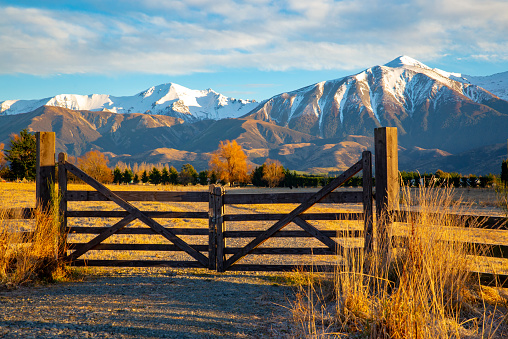 A closed wooden farm gate at the entrance to a farm field below snowy mountains on a frosty winter morning.
