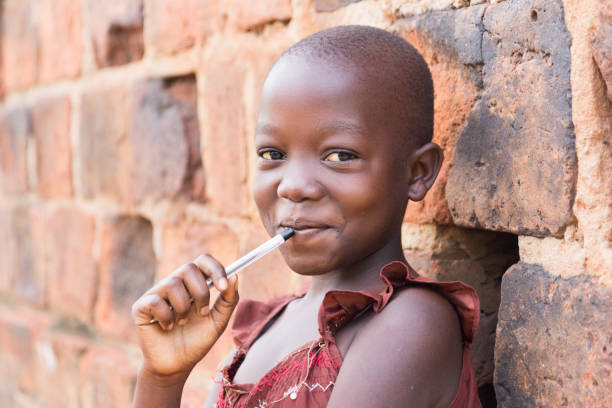 a smiling Ugandan girl An 11-year-old Ugandan girl smiling, holding a pen against her mouth and leaning against a brick wall looking at the camera uganda stock pictures, royalty-free photos & images
