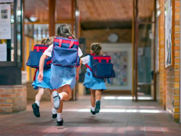 Rear view of excited students running towards entrance. Girls are carrying backpacks while leaving from school. Happy friends are wearing school uniforms.