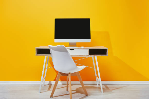 istock Computer with blank screen at wooden table with chair near yellow wall 1071308842