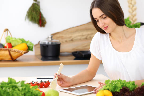 istock Beautiful Hispanic woman cooking in kitchen while using tablet computer and wooden spoon. Housewife found new recipe for dinner or breakfast. Healthy meal and householding concepts 1071315616