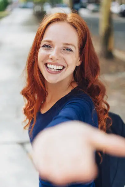 Vivacious happy young woman with long red hair holding out her hand to the camera with a beaming toothy smile