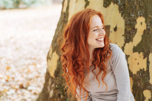 souriante jeune femme appuyée contre un tronc d’arbre - women autumn beauty in nature smiling photos et images de collection
