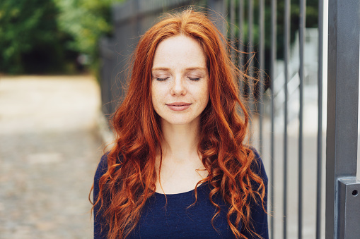 Serene young redhead woman standing with closed eyes outdoors in a park alongside an iron railing