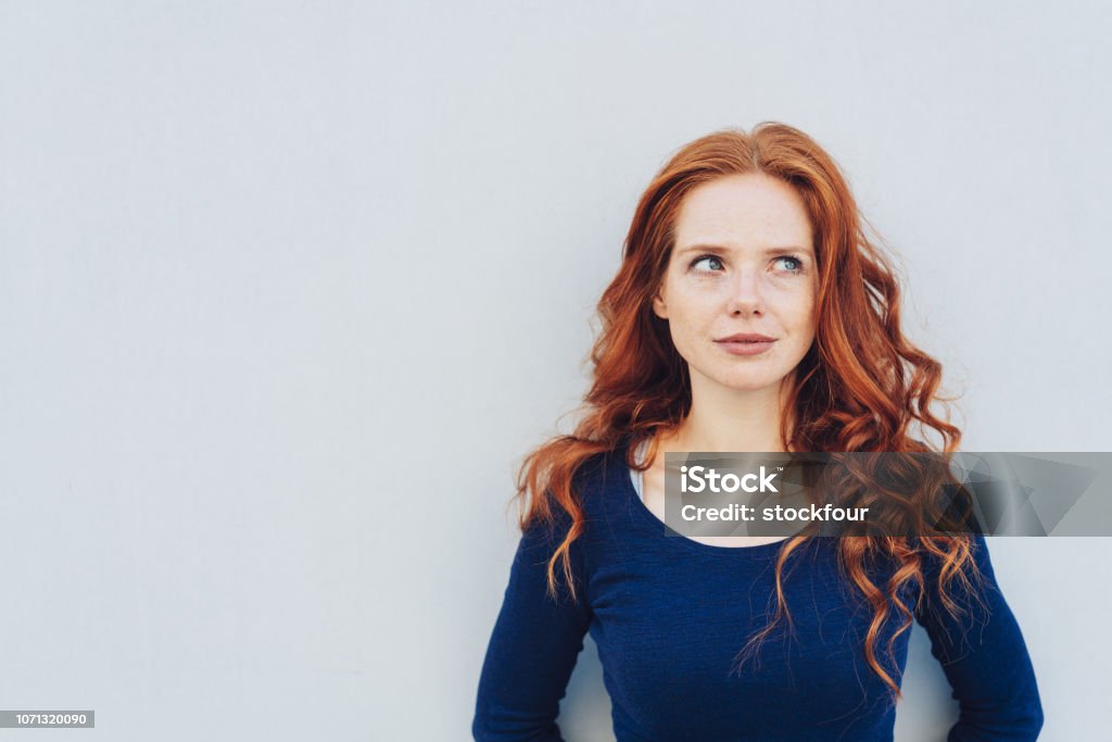 Attractive young woman standing pondering Attractive young woman standing pondering a problem looking up with a contemplative expression against a white exterior wall with copy space Women Stock Photo