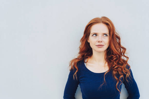 Attractive young woman standing pondering Attractive young woman standing pondering a problem looking up with a contemplative expression against a white exterior wall with copy space redhead stock pictures, royalty-free photos & images