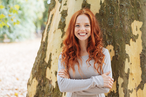 Smiling confident young woman with lovely long curly red hair standing with folded arms in front of a tree trunk in a park