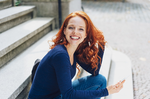 Young cheerful red-haired woman sitting on steps with mobile phone
