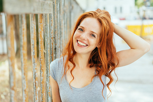 attractive friendly woman with tousled long red hair standing against a metal railing in a high key outdoor street