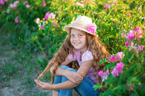 Cute brunette child blue eyes girl is holding flowers standing in pink rose field. Wearing stylish dress and hat, enjoy summer time days childhood.