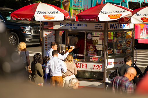 Some tourists are buying hot dogs from a kiosk in Times Square. Times Square is a major commercial intersection, tourist destination, entertainment center and neighborhood in the Midtown Manhattan section of New York City.