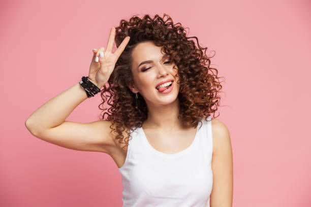 imagem de feliz jovem pé isolado sobre fundo rosa mostrando o gesto de paz. olhando a câmera - cabelo encaracolado - fotografias e filmes do acervo