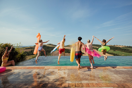 Rear view of friends jumping in the air ready to splash into the pool. They're all carrying an inflatable each.