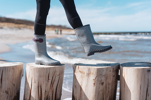 Woman wearing gumboots walking along wooden poles used as a coastal barrier on a sandy beach with waves in a low angle view of her lower legs