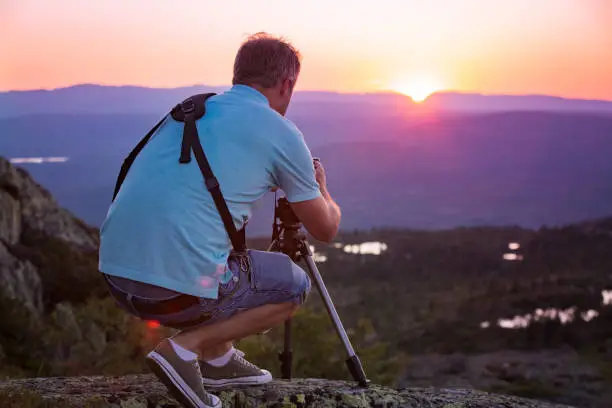 Portrait of man with camera and tripod filming sunset above mountain. Traveler Taking pictures from the top of mountain. Beautiful view of pink sky, Norway, Kongsberg