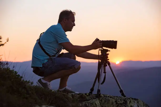 Portrait of man with camera and tripod filming sunset above mountain. Traveler Taking pictures from the top of mountain. Beautiful view of pink sky, Norway, Kongsberg