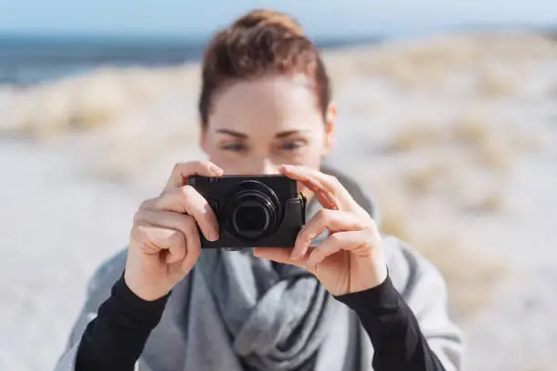 Attractive woman tourist taking photos on a beach in a warm woolly outfit looking at the viewfinder to check the image with focus to the camera