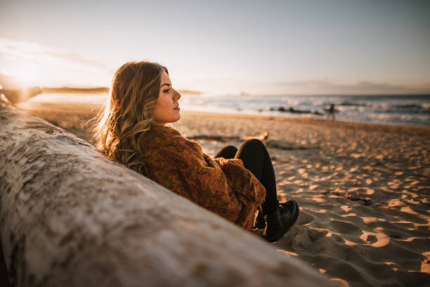 jeune femme assise sur une plage au coucher du soleil en hiver - beach nature outdoors overcast photos et images de collection