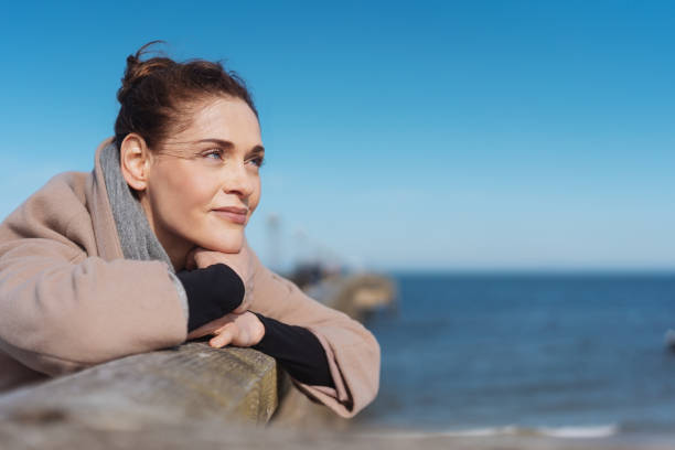 Pensive young woman relaxing on a wooden pier Pensive young woman relaxing on a wooden pier enjoying the winter sunshine leaning on the railing staring out over the ocean mid adult women stock pictures, royalty-free photos & images