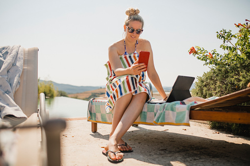 Mid-adult female working by the side of the pool. She is sitting on a sun lounger and using her laptop to reply to emails.