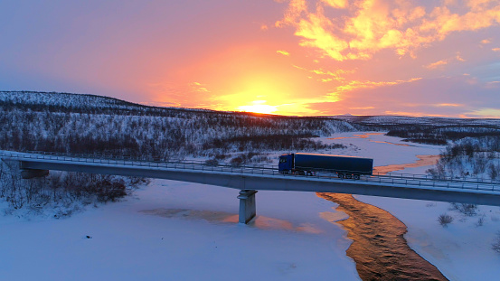 AERIAL: Semi truck crossing the bridge above icy river in the winter at sunset