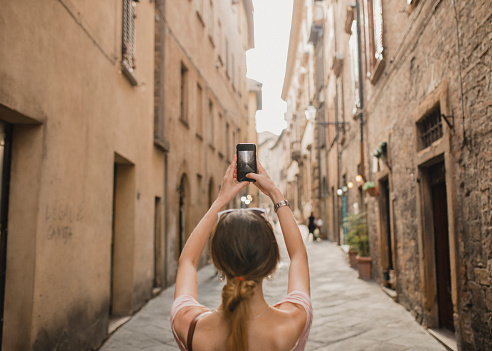 Rear View of an unrecognisable person standing in a street in the old town of Volterra. She is taking a photo of the architecture within the street.
