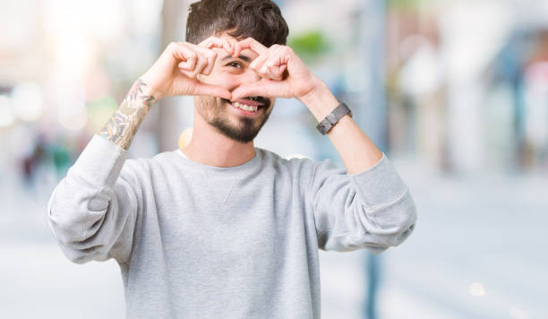 young handsome man wearing sweatshirt over isolated background doing heart shape with hand and fingers smiling looking through sign - made man object imagens e fotografias de stock