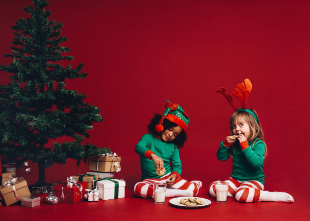 Two little girls dressed in christmas theme clothes sitting beside a christmas tree. Happy kids sitting beside a christmas tree enjoying cookies with milk.