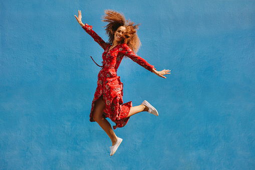 Young female model in red sundress jumping with joy. Beautiful young woman jumping over blue background.