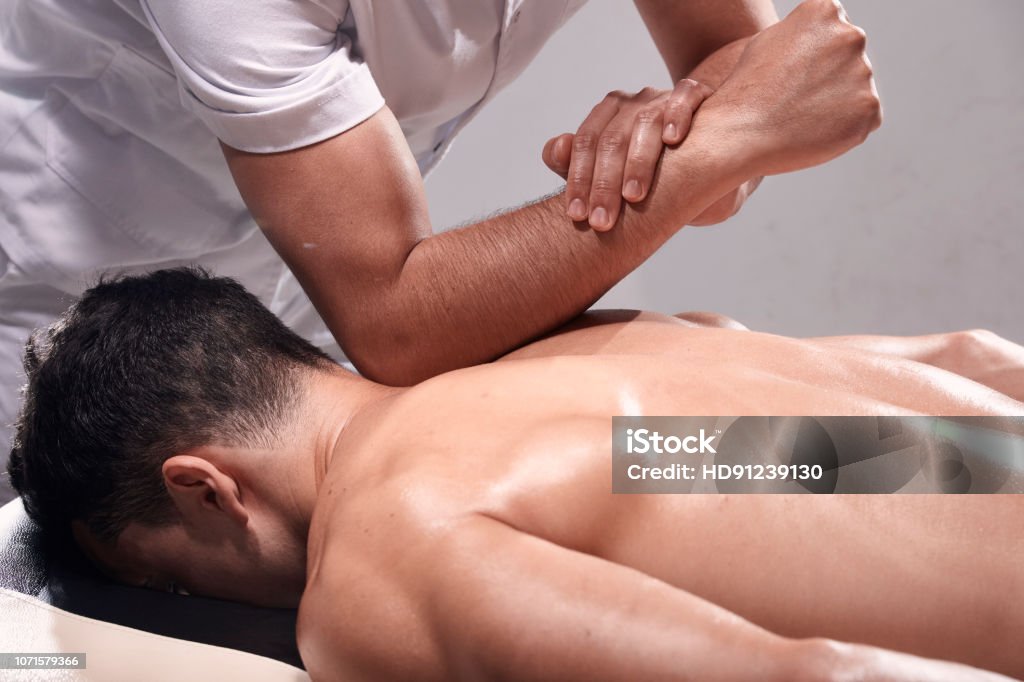 side view, two young man, 20-29 years old, sports physiotherapy indoors in studio, photo shoot. Strong Physiotherapist hard massaging relaxed patient neck back side, with his elbow. Massaging Stock Photo