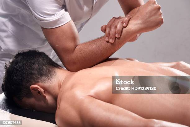 Side View Two Young Man 2029 Years Old Sports Physiotherapy Indoors In Studio Photo Shoot Strong Physiotherapist Hard Massaging Relaxed Patient Neck Back Side With His Elbow Stock Photo - Download Image Now