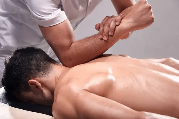 Photo of side view, two young man, 20-29 years old, sports physiotherapy indoors in studio, photo shoot. Strong Physiotherapist hard massaging relaxed patient neck back side, with his elbow.