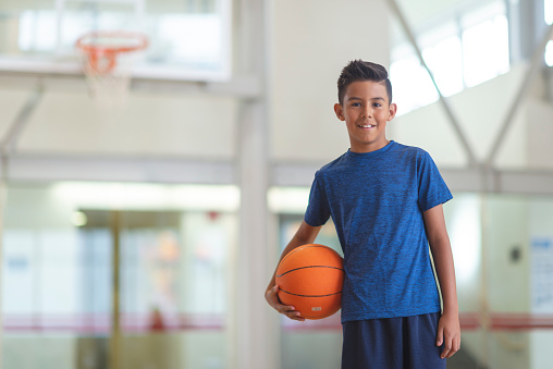 A boy is standing in a gymnasium holding a basketball. He is smiling at the camera.