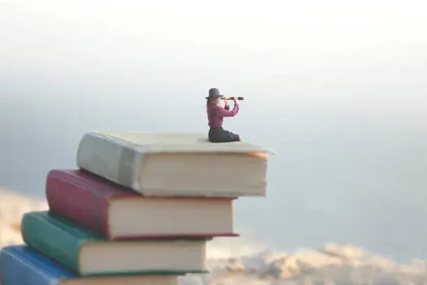 Photo of miniature woman looks at the infinity with the spyglass on a scale of books
