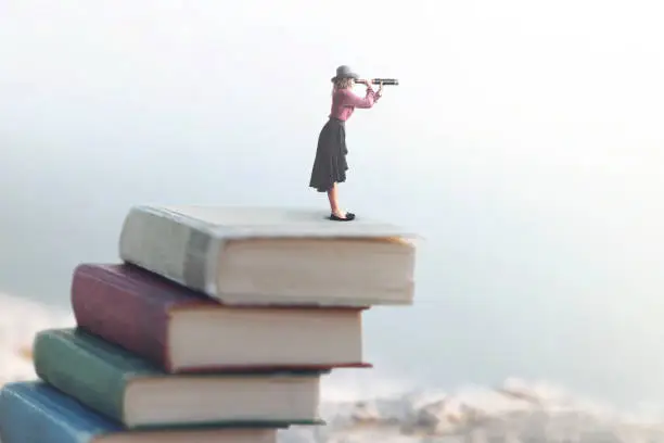 Photo of miniature woman looks at the infinity with the spyglass on a scale of books