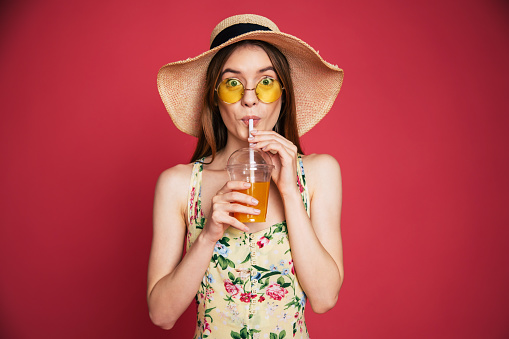 Portrair of happy and excited blond trendy woman in hat with fresh drink in hands isolated on pink background. Summer mood
