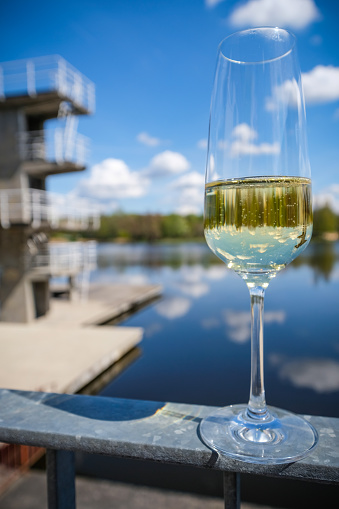 Close-up on a glass of sparkling champagne mirroring the diving platform, blue sky and clouds and the lake in the background