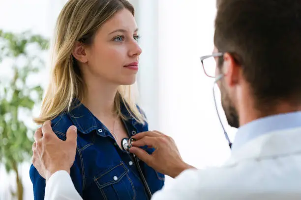 Shot of handsome young male doctor checking beautiful young woman patient heartbeat using stethoscope in medical office.