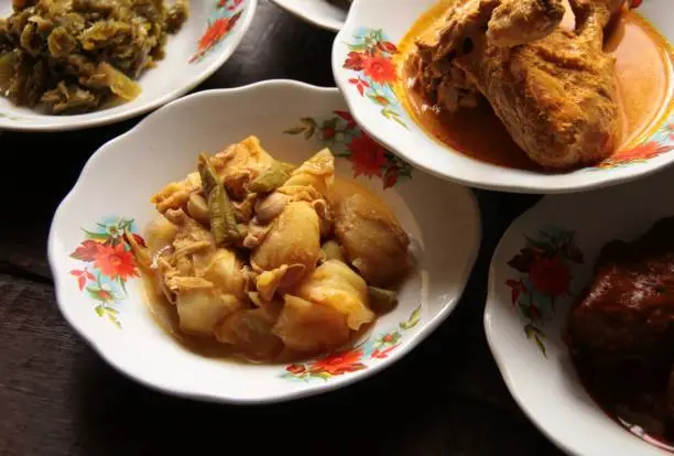 Sayur Gulai Nangka, the popular vegetable dish of jackfruit in yellow curry. Plated in a small ceramic plate and served among many other Minang side dishes in style typical to Minang eateries, plates stacking.